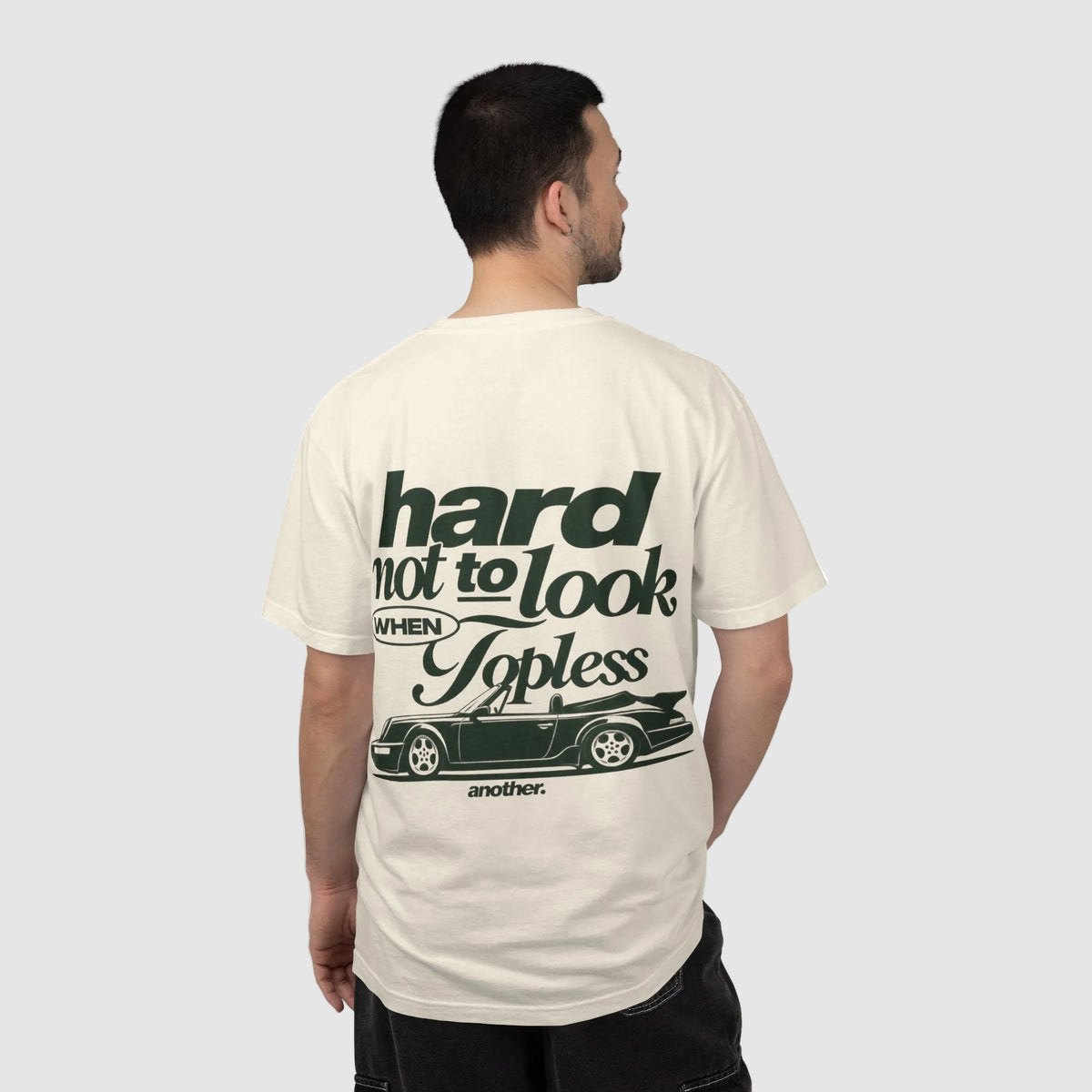 A man viewed from behind wearing a cream-colored t-shirt with a dark green graphic print on the back. The graphic features the text 'hard not to look WHEN Topless' above a silhouette of a classic car, with 'another.