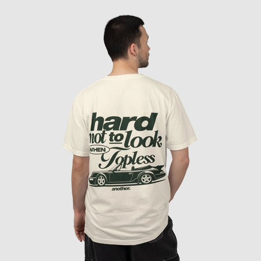 A man viewed from behind wearing a cream-colored t-shirt with a dark green graphic print on the back. The graphic features the text 'hard not to look WHEN Topless' above a silhouette of a classic car, with 'another.