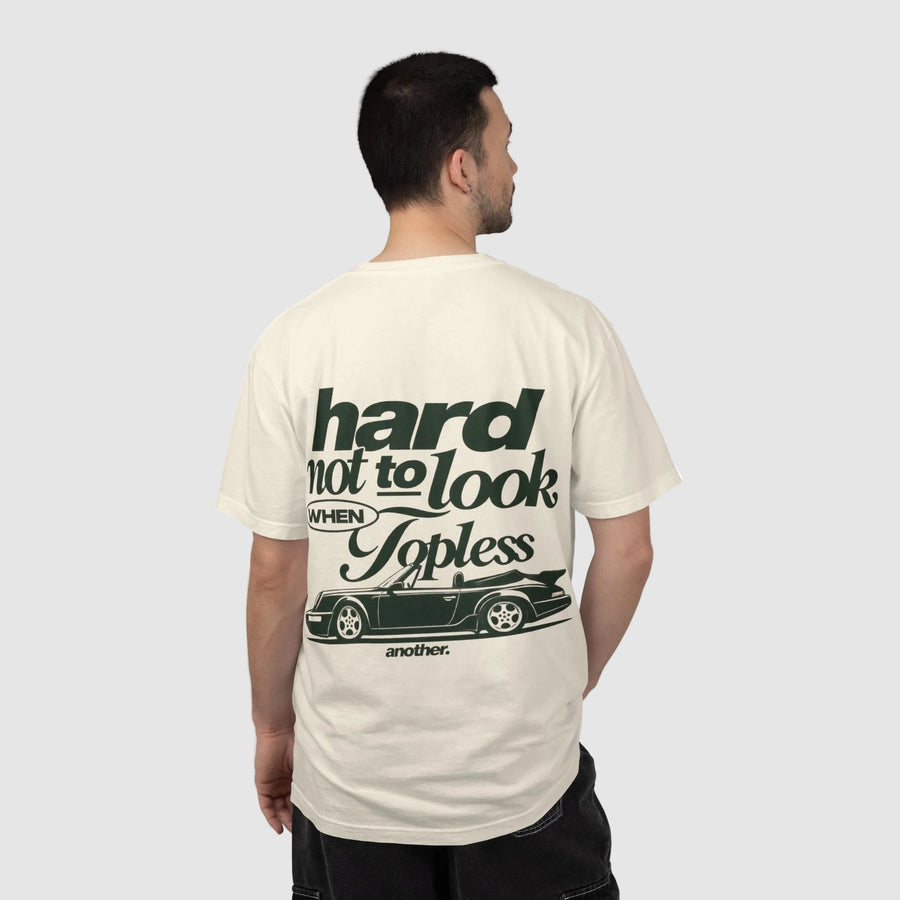 A man viewed from behind wearing a cream-colored t-shirt with a dark green graphic print on the back. The graphic features the text 'hard not to look WHEN Topless' above a silhouette of a classic car, with 'another.