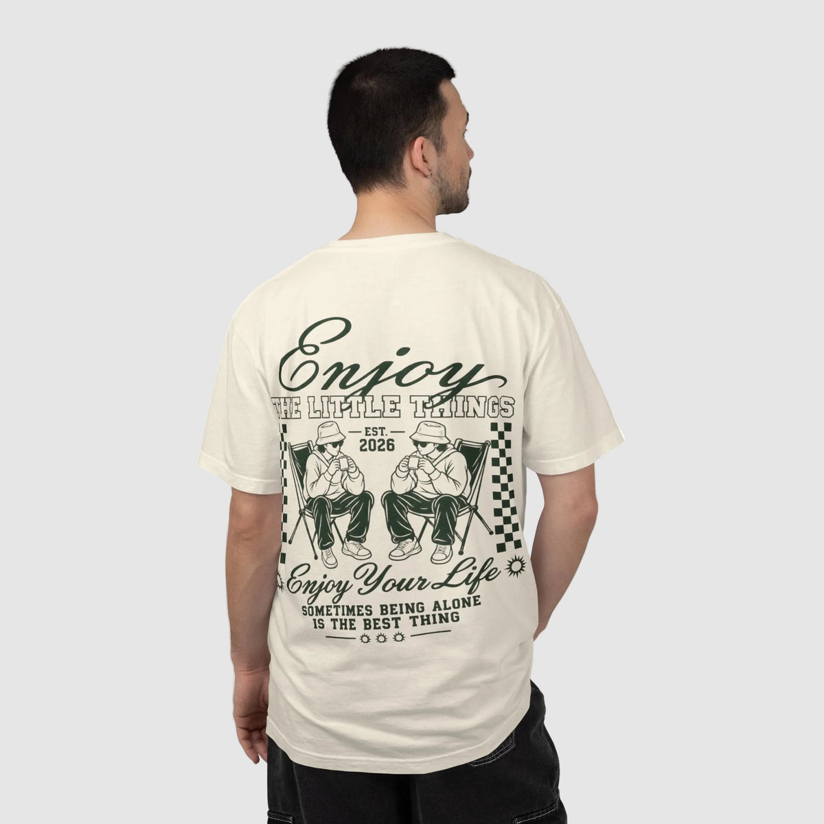 A man shown from the back wearing a cream-colored t-shirt with a large dark green graphic print on the back. The graphic features stylized illustrations of two men in hats and the text 'Enjoy The Simple Things' and 'Enjoy Your Life'.
