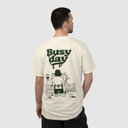 A man shown from the back wearing a cream-colored t-shirt with a dark green graphic print that reads 'Busy day' above an illustration of a person working at a coffee station.