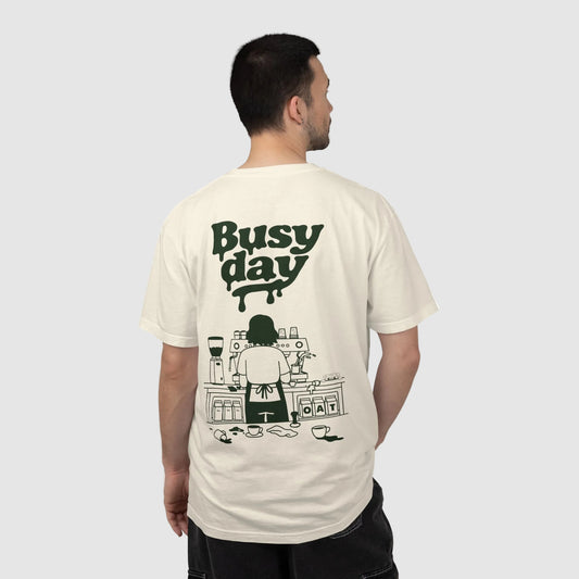 A man shown from the back wearing a cream-colored t-shirt with a dark green graphic print that reads 'Busy day' above an illustration of a person working at a coffee station.