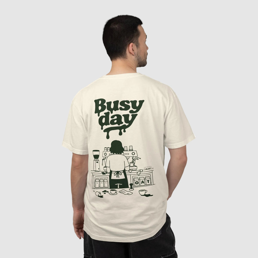 A man shown from the back wearing a cream-colored t-shirt with a dark green graphic print that reads 'Busy day' above an illustration of a person working at a coffee station.
