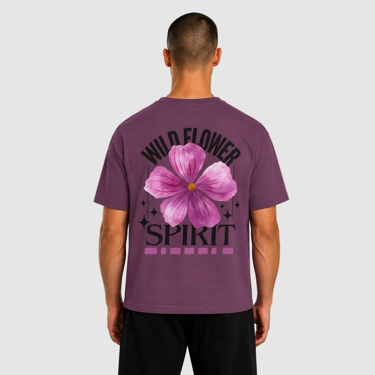 A man viewed from the back wearing a purple short-sleeve t-shirt with a large graphic on the back. The graphic features a pink hibiscus flower in the center, with the words 'WILD FLOWER SPIRIT' in black arched above and below the flower.